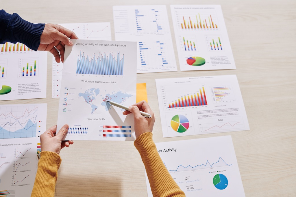 View from above of desk covered in charts and graphs with 2 people's hands holding them and pointing at contents